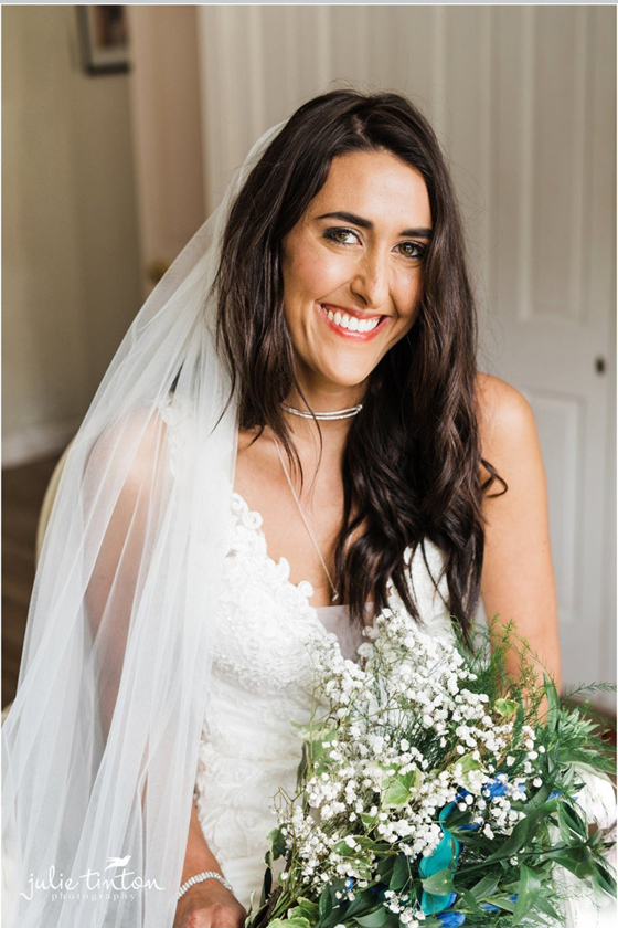 Bride smiles in hotel room with bouquet in hand and veil draped over her shoulder, her makeup is done by Jennifer Louise Studio.